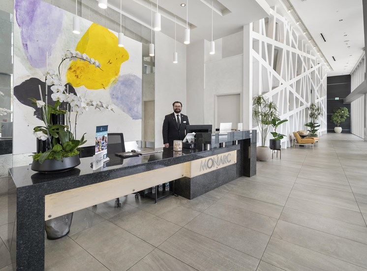 A man in a suit stands behind a reception desk with a large yellow flower on the wall behind him.